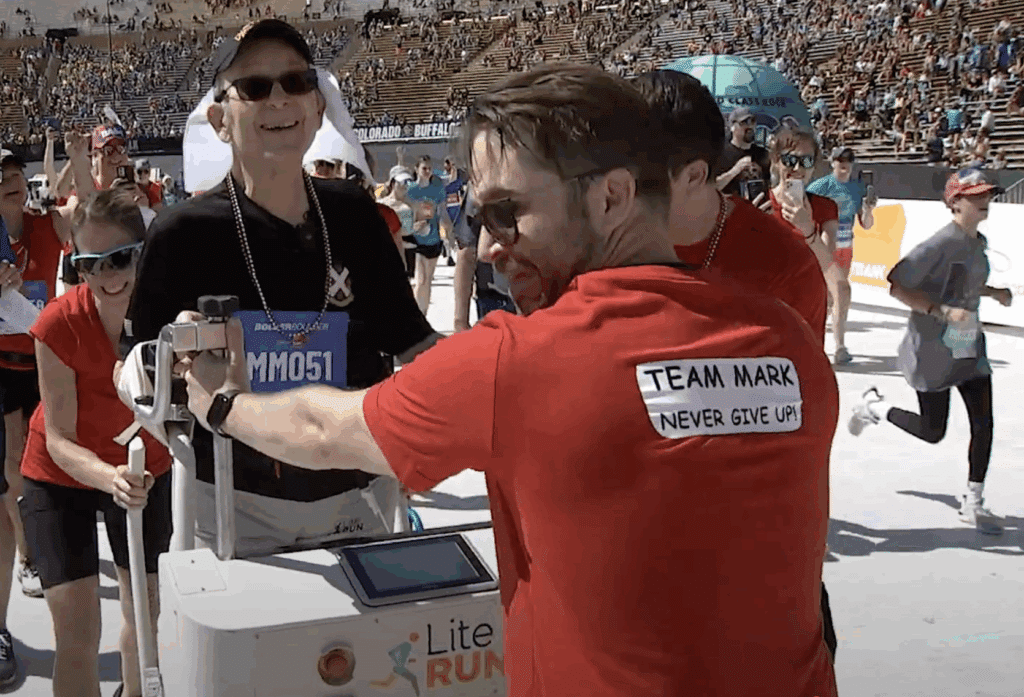 A man using a Lite Run Mobility Trainer in a stadium at the finish line of a race. There are other runners and people helping the man also in the frame. One man is wearing a red shirt with "Team Mark Never Give Up" written on the back.
